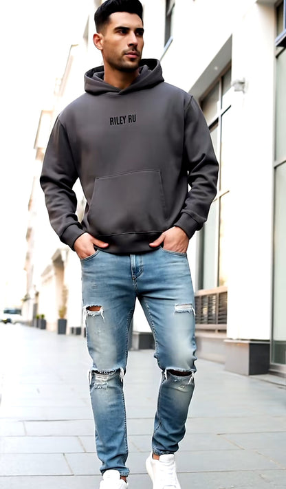 Man wearing a dark hoodie and ripped jeans standing on a city street.