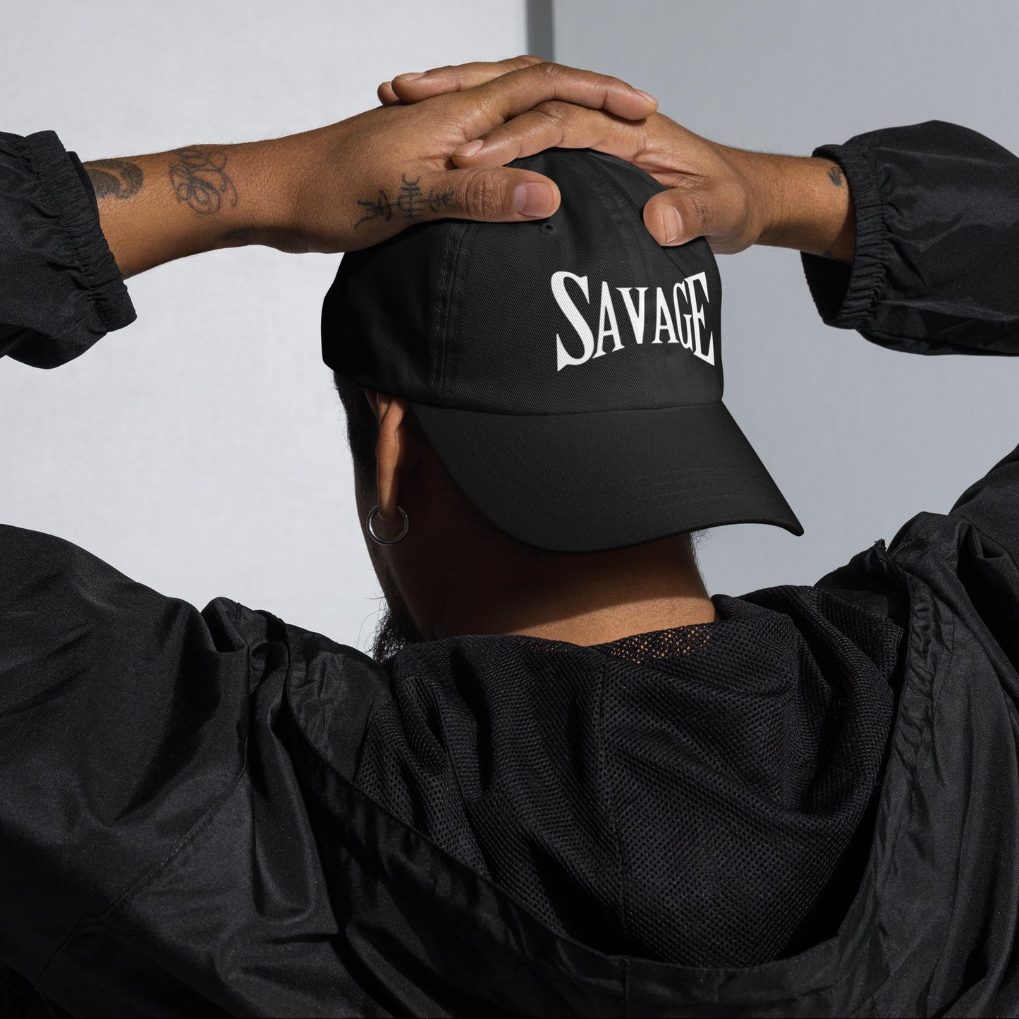 Person wearing a black cap with 'SAVAGE' hands on head, against a neutral background