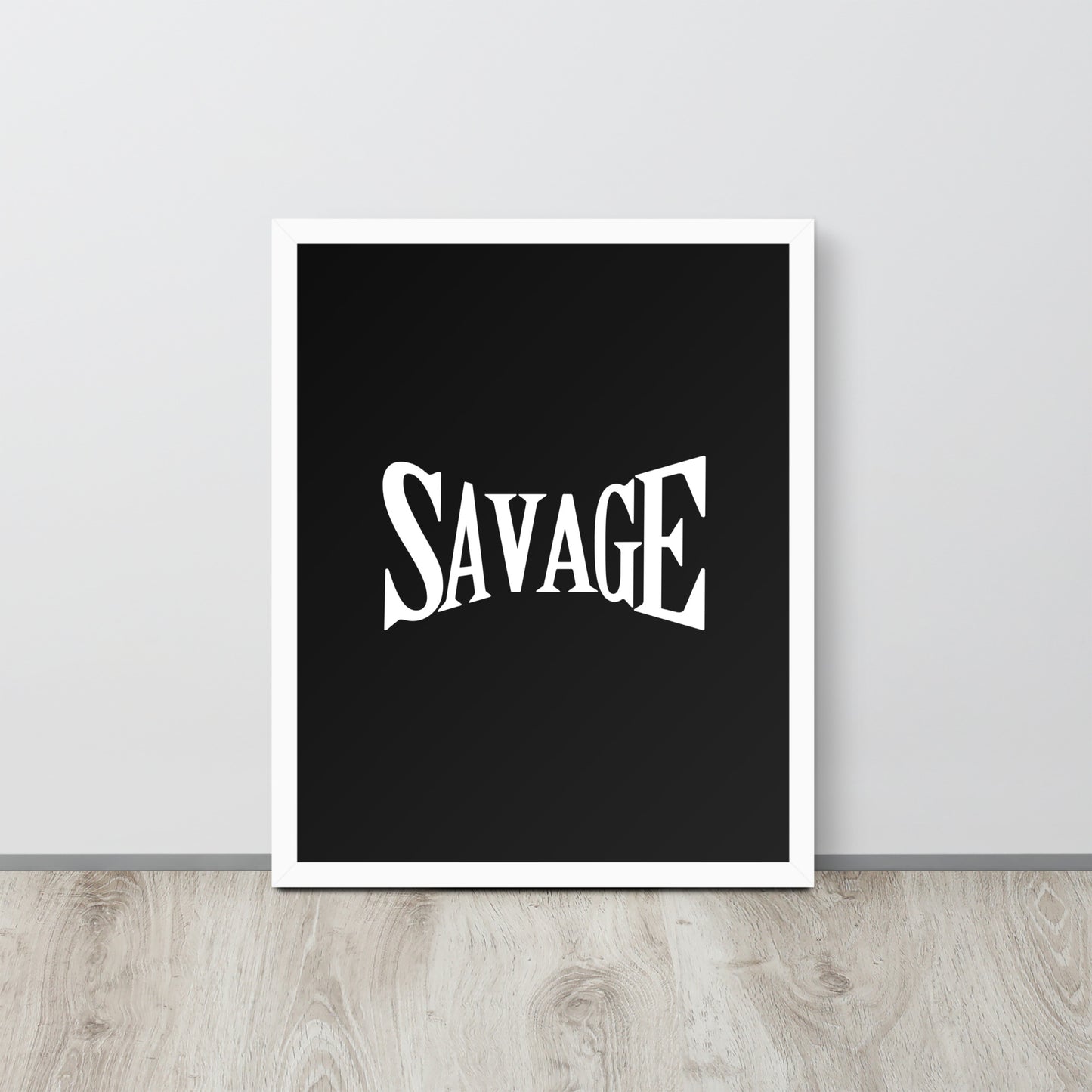 Framed poster with the word 'SAVAGE' on a black background, placed on a wooden surface with a light gray wall in the background.