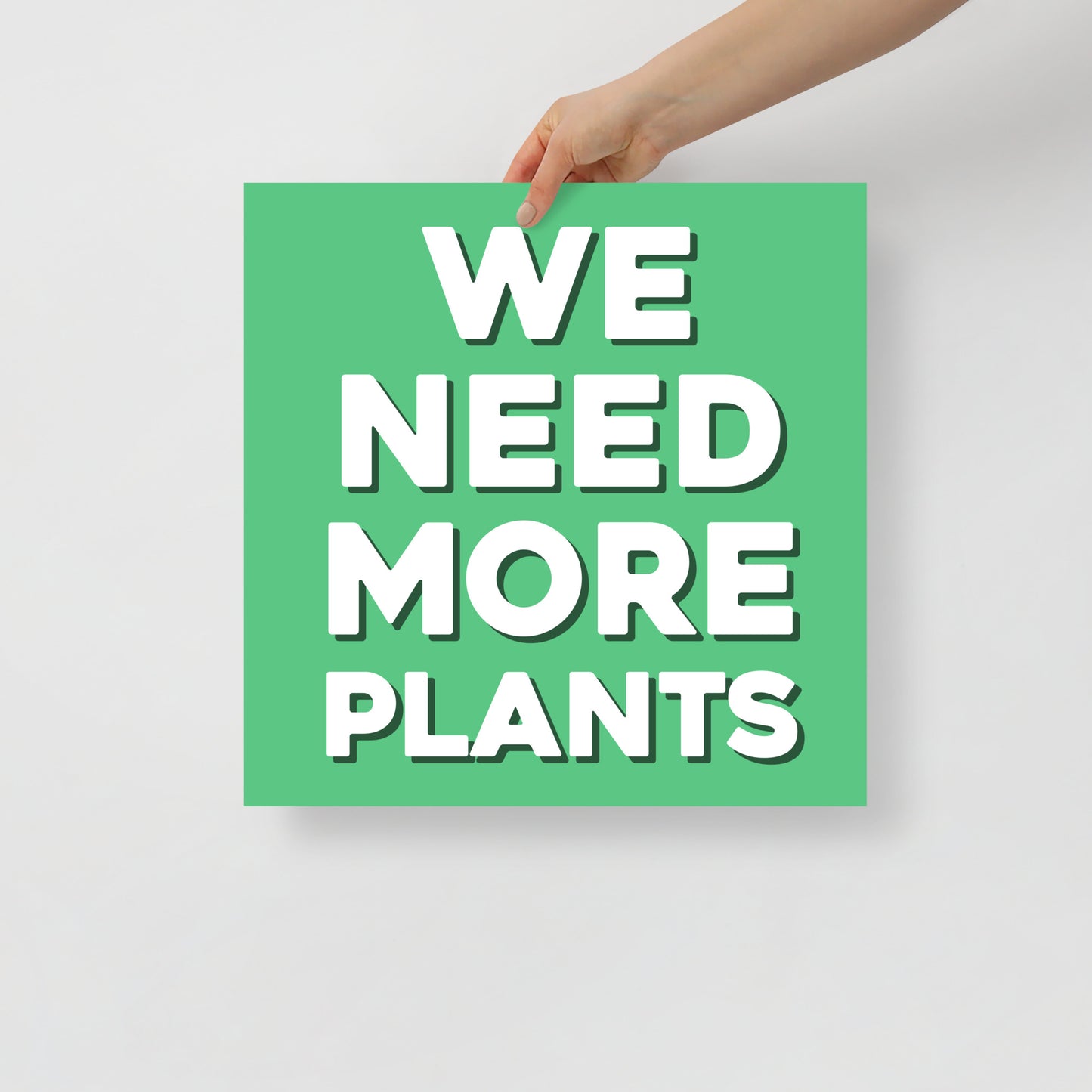 Hand holding a green sign with 'WE NEED MORE PLANTS' text on a light gray background