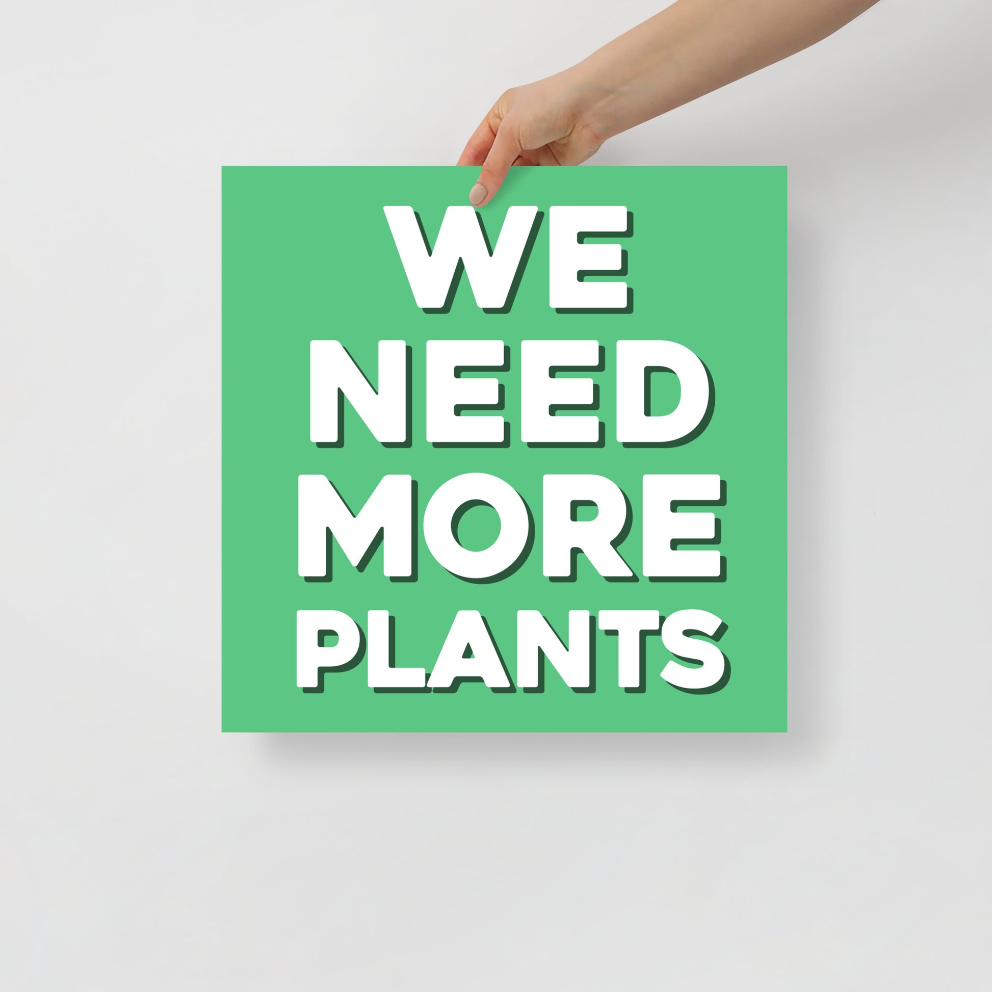 Hand holding a green sign with 'WE NEED MORE PLANTS' text on a light gray background