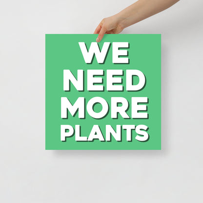 Hand holding a green sign with 'WE NEED MORE PLANTS' text on a light gray background