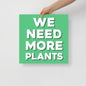 Hand holding a green sign with 'WE NEED MORE PLANTS' text on a light gray background