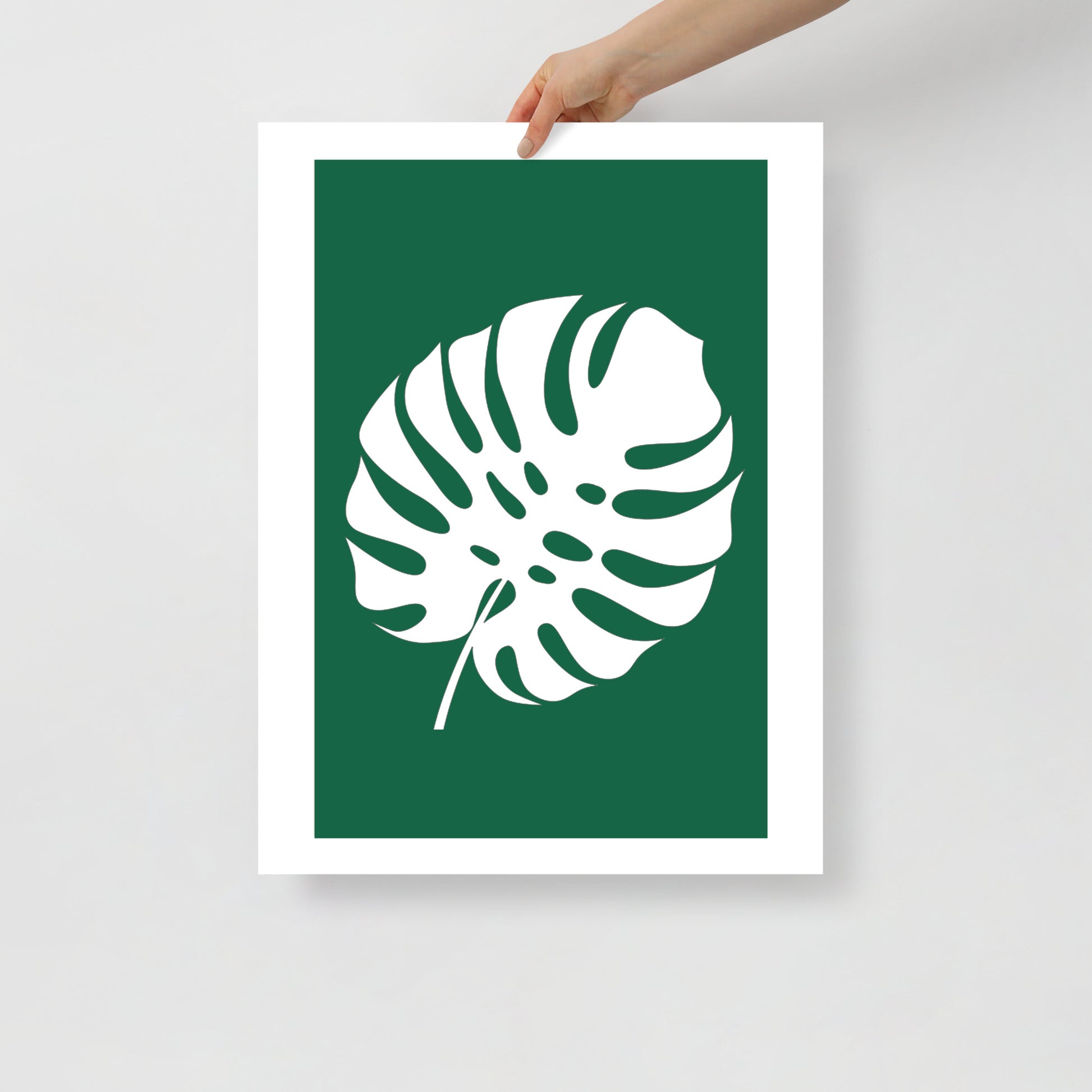 Framed poster with a white leaf design on a green background, held by a hand against a light gray background.